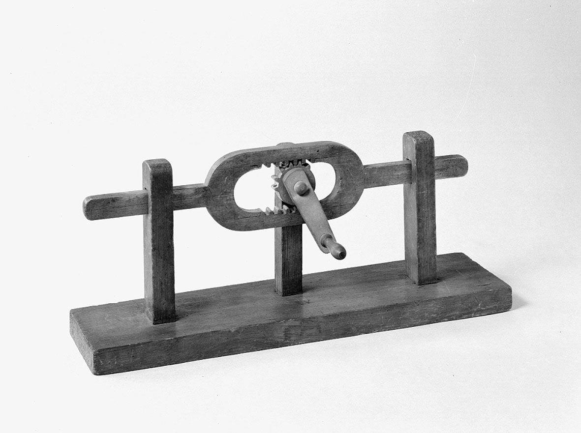 Modell ur Polhems mekaniska alfabet. Text på föremålet: "XV". En roterande rörelse hos hjulet, som till hälften är försett med kuggar, överförs till en fram- och återgående rörelse hos den bygelförsedda armen.