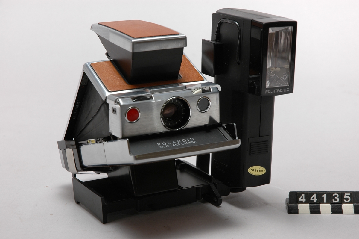 Direktbildskamera Polaroid Land Camera SX-70 med väska, blixthållare, stativhållare, trådutlösare samt 25 st filter. Kromplastad SLR kamera (Single-Lens Reflex) med påklistrad läderdekoration, 4-elements 116 mm f/8-f/22 glaslins och elektroniskt styrd slutare med hastigheter från 1/175 upp till 10 sekunder. På kamerans front finns avtryckarknappen, exponeringsmätarens sensorfönster, reglage för skärpeinställning och kompensering av den automatiska exponeringsinställningen och en sockel för att ansluta en modul med 10 st blixtlampor. Manuellt fokus kan ställas in ner till 26,4 cm. SX-70 var den första SLR-kameran på marknaden för direktframkallade bilder och den första som använde Polaroid's  nya integrerade färgfilm som framkallades i fullt dagsljus utan manuella ingrepp av fotografen. Filmpacken, som bestod av 10 bilder med formatet 7,9 x 7,9 cm, innehöll dessutom ett tunt platt 6-volts batteri av zink-klorid som försörjde exponeringselektroniken, motorn till filmframmatningen och blixtlamporna. Filmens hastighet var ASA 150. Kameran kan fällas ihop till ett format som får plats i en kavajficka.
Tillbehör: Väska, blixthållare, stativhållare, två st. filter och trådutlösare.