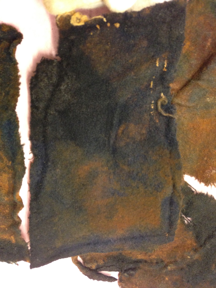 Textil. 13 tygfragment uppdelade på fyndnummer 03466a-d.
Fnr 03466a består av 10 fragment av ull vävda i tuskaft varav det största är ihopsytt av flera delar. Har varit ett par byxor med lucka fram. Originalfärgen kan ha varit blå då rester av blå färg syns på insidan av två av bitarna på de delar där sömmen varit och tyget originalyta bevarats bättre. På en av dessa bitar som tolkats som en del av en ficka på byxans vänstra sida finns rester av en tvåtrådig, s-tvinnad, z-spunnen tråd i sömmen. Byxans tyg har varit mer valkad på insidan än på utsidan. På det största fragmentet framsida syns två knapphål i linningen, och rester efter ytterligare två hål. Det kan därför konstateras att det ursprungligen har varit två knapphål på var sida på linningens mittdel där sidodelarna av linningen knäppts eller knutits fast. Vid ena knapphålet syns något som kan vara resterna av en knapp eller hyska odyl. När byxans delar läggs ihop som de en gång suttit mäter midjebredden ca 47 cm vilket ger ett midjeomfång på ca 94 cm före ihopsnörning. Av det som kvarstår av byxbenen kan konstateras att dagens (år 2015) längd på vänster byxben är 60 cm från linningens övre kant och ner. Höger byxben är i dagsläget endast 43 cm långt. 
På fnr 03466b syns rester av söm i en av kanterna. Fragmentet är av ull och vävt i 2/2-kypert. Lappen har en söm i en cirkel mitt på tyget som troligen är resterna efter den söm som sytt fast lappen runt ett hål på byxan. Kan ha varit en lagningslapp på byxans högra knä som uppvisar motsvarande söm kring ett cirkelformat hål. 
Fnr 03466c är ett fragment av ull vävt i lite finare tuskaftsväv än fnr 03466a. Fragmentet har hål efter sömmar i kanterna samt delar av en ljus tråd i en av kanterna. Blå färg syns där tyget varit skyddat där sömmen varit. Fragmentet kan ha varit en innerficka bak på byxans vänstra sida. Det finns en söm i byxans tyg där som överensstämmer med lappens söm.
Alla kanterna på fnr 03466d är invikta och har hål efter söm som om biten varit fastsydd på ett annat tygstycke. Fragmentet är av ull och vävt i samma sorts tuskaftsväv som byxan. Kan ha varit en lagningslapp på byxan över ett hål i byxtyget på den plats där vänster innerficka bak är placerad.

Tidigare beskrivning: Stort tygskynke. Beckigt.