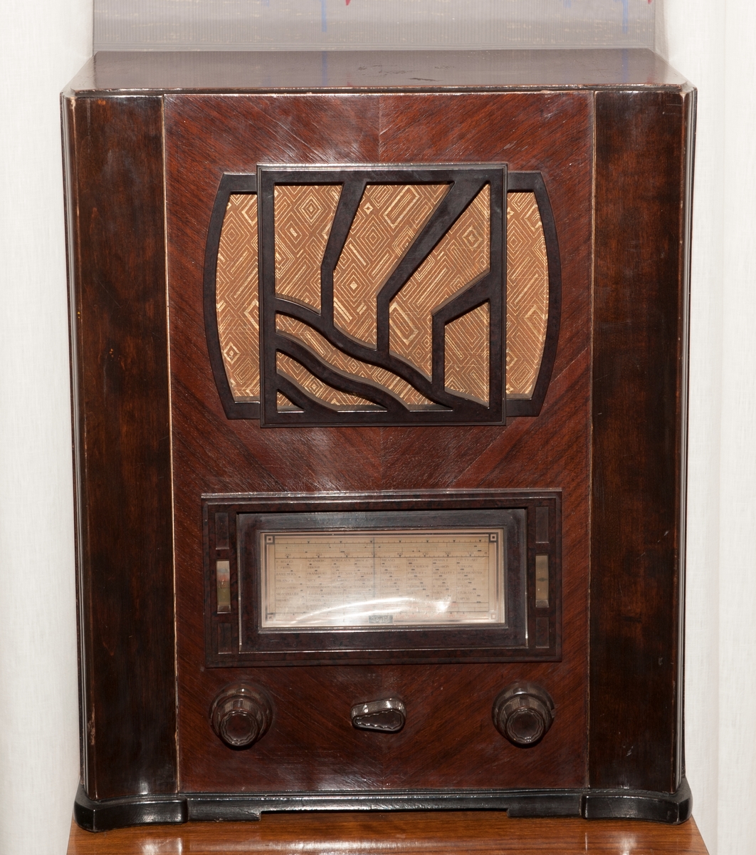 Radiokabinett i brunt tre med mørkebrunt listverk. 
3 innstillingsknapper og  Art-Deco-dekor på front. 