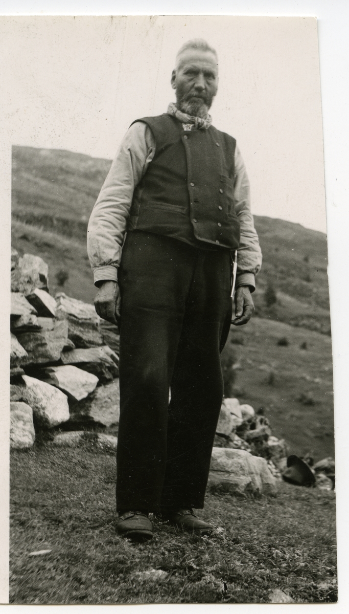 Portrett i helfigur av en mann som står foran en steinmur. Mannen er iført vest, mørke bukser og et halstørkle. Hatten ligger på bakken i bakgrunnen.