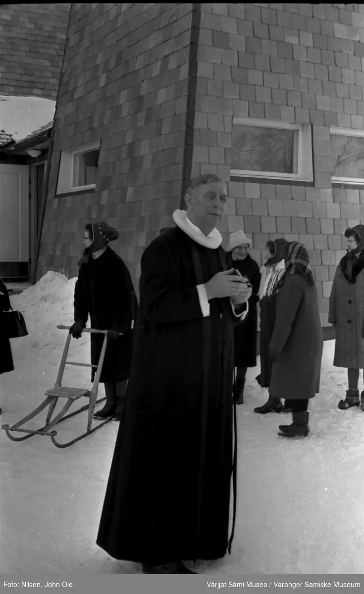 En prest står utenfor det som sannsynligvis er Tana kirke i Rustefjelbma. En gruppe kvinner står i bakgrunnen. 1967.