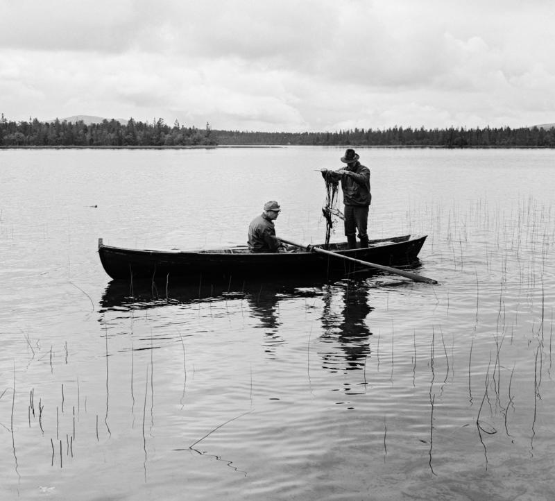Two men in a rowing boat. One man rows and the other fishes.