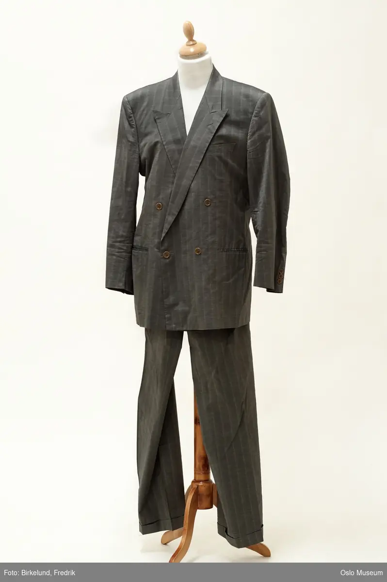 Skreddersydd dress fra 1980-tallet i grått, blankt stoff med striper. Dobbeltspent jakke med skulderputer. Bukse med vide ben og legg i livet.