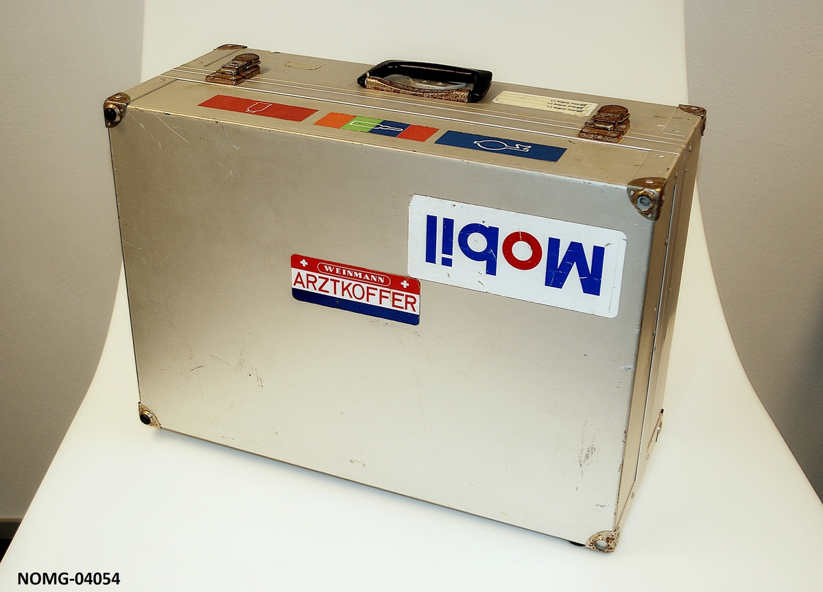 Legekoffert med hank og lås. Skulderrem på den ene siden. Tre rom inni med diverse medisinske utstyr.