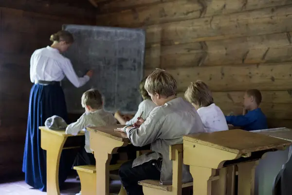 Children at their desks in the old school house while teacher writes at the black board