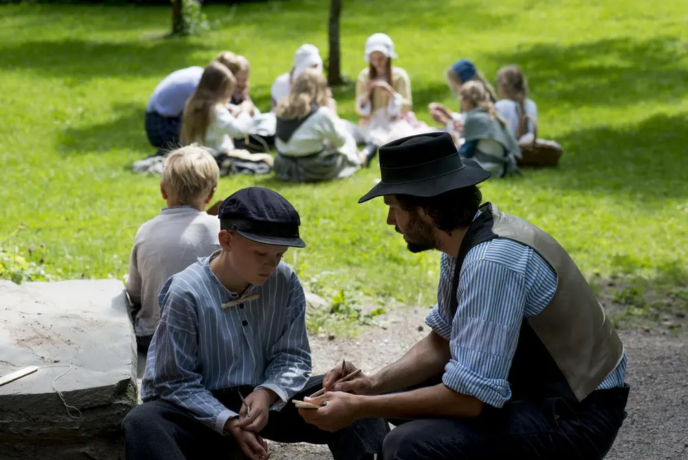 En ferieskolegutt og drengen i forgrunnen. Andre ferieskolebarn sitter i en ring på gresset i bakgrunnen