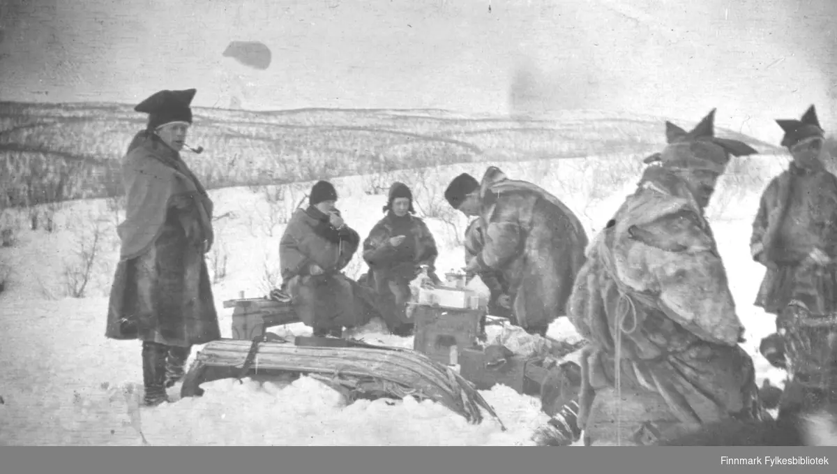 Aimjok,1916. Samer i pesk med karakteristisk samisk lue. En av mennene røyker pipe. Noen sitter med mat eller drikke i hendene.