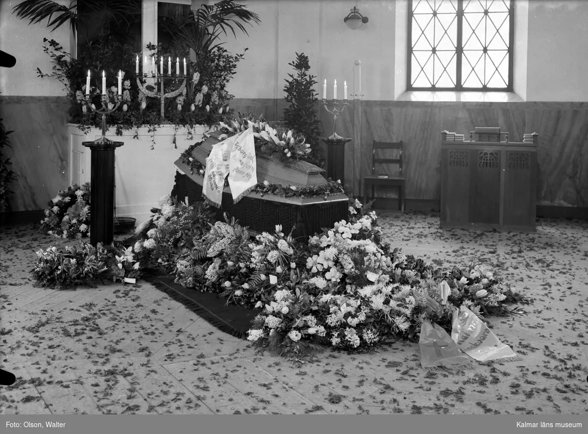 Kista framför altaret med blommor och kransar. Beställare: Bergs Begravningsbyrå, Kalmar.