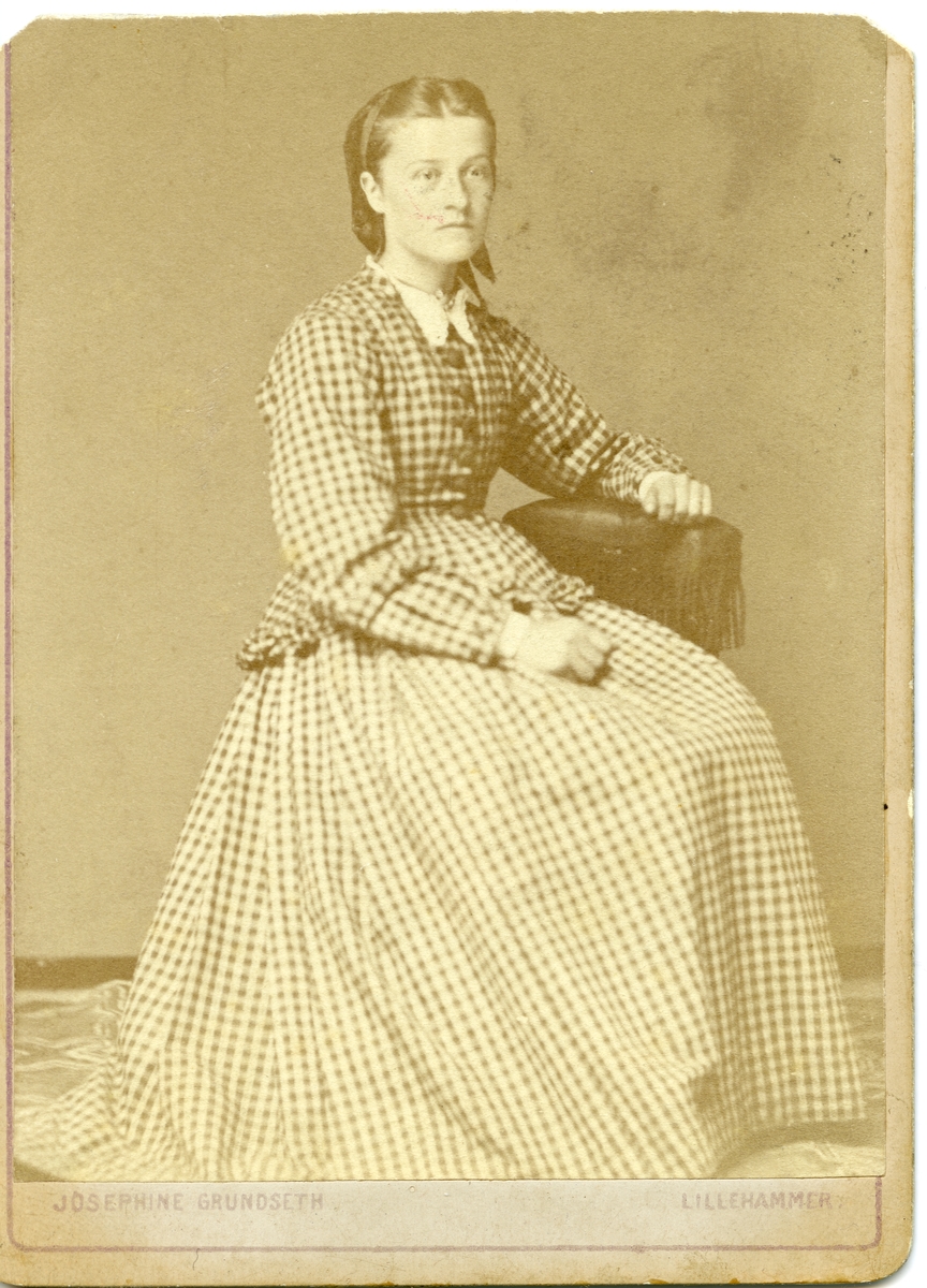 Portrett av en kvinne sittende på en stol. Kvinnen er iført en rutete kjole.
