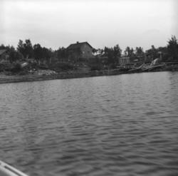 Bilde tatt fra innsjøen mot et hus og noen hytter langs bred
