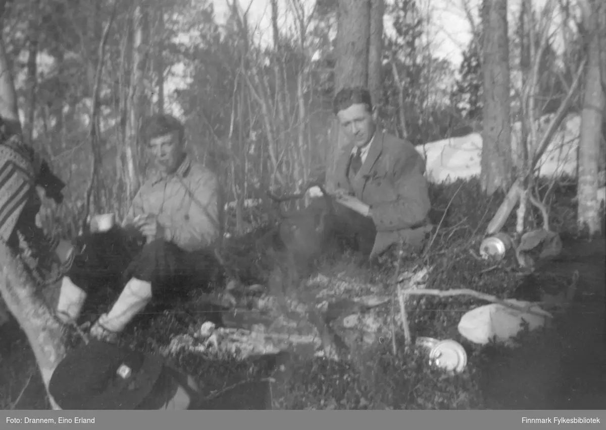 To menn sitter ved et lite bål i skogen. Sted og personer er ukjent.