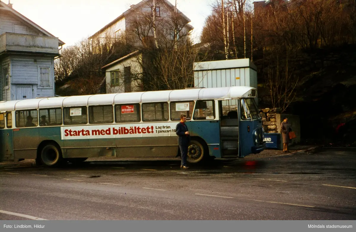 En man och en kvinna står vid en blå buss på en busshållplats vid Gamla Torget i Mölndal, 1970-tal. Till vänster ses del av huset Kvarnbygatan 37.

För mer information om bilden se under tilläggsinformation.