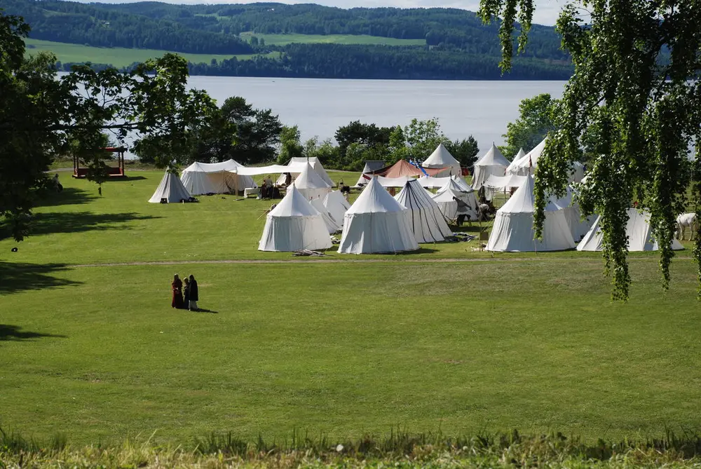 Leir av hvite middelaldertelt står på ei stor gresslette omkranset av løvtrær og med Mjøsa i bakgrunnen.