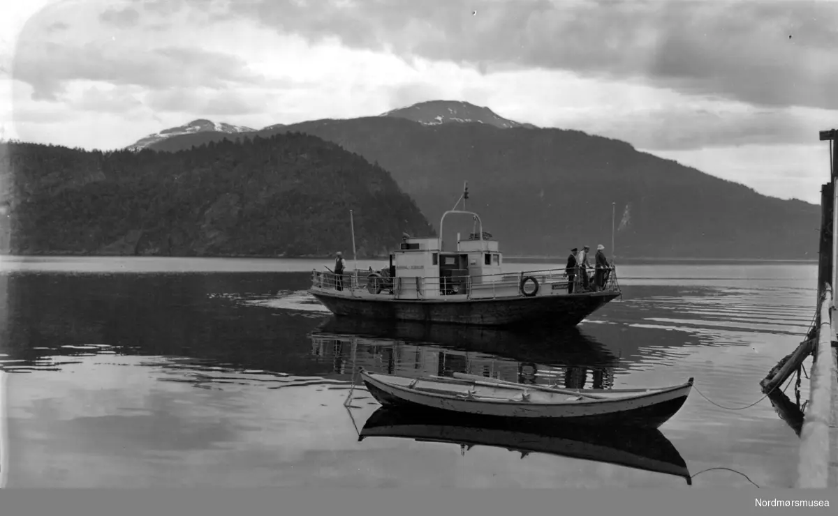 En bilferge på vei mot kai. En robåt ligger i forkant. serie Fra Nordmøre museums fotosamlinger.

