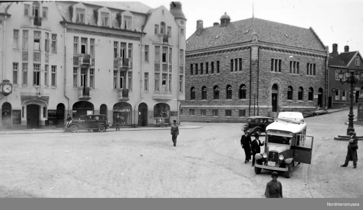 Torget på Kirkelandet i Kristiansund. Grand hotell til venstre, og Norges Bank til høyre i bildet.  - Rutebil til høyre med registreringsnummer T-4274 står i Norges Bilbok 1935 på Studebaker 1932, Kristiansund Oppdal Auto. Bilen bak er den nyeste, 1933-34-modell, med tanke på datering av bildet. Den foran hotellet er typisk ca. 1930. (info: Ivar Stav). Kommentar: 
T-4274 var en buss med 18 seter, 1932-modell, en stor buss for sin tid. 1932 var en milepæl i rutebiltrafikken i regionen. Den våren ble det satt inn bilferje Kvisvik-Kristiansund, og rutebilene fra Oppdal kunne følge ferja helt fram til Kristiansund sentrum. Fram til da var det båt den siste strekningen til byen. Dette var raskeste vei mellom Kristiansund og Oslo, med bytte til tog på Oppdal stasjon. Sundal-Opdal Auto skiftet samtidig navn til Kristiansund-Opdal Auto (KOA), et navn som var i bruk i 50 år, til 1982 (dobbel P i Oppdal kom etter krigen).  (info:
Sveinung Berild).  Bildet er trolig fra juni 1933 ut fra forretningen Astrid Giertsen holder sluttsalg i Grand Hotell. Fra Nordmøre museums fotosamlinger.