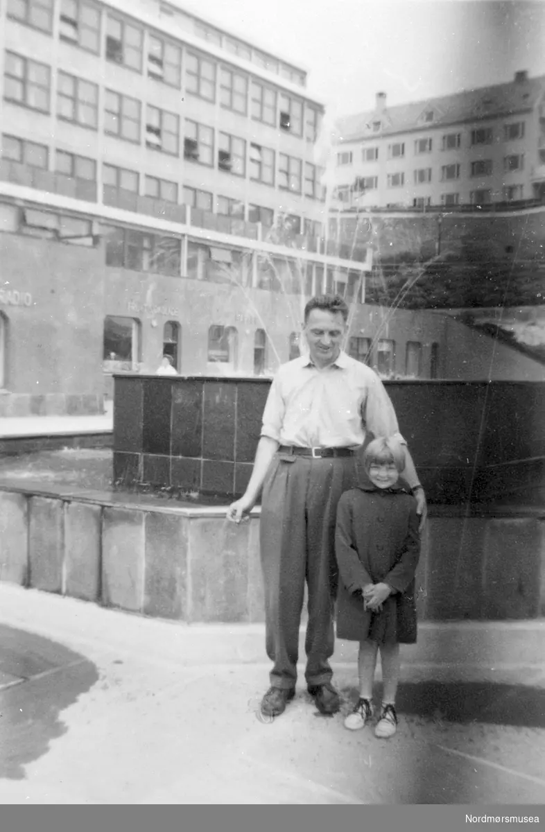 Kristiansund, Rådhusplassen utpå 1950-tallet. Far og datter foran fontena. Som vanlig er det vannsøl. Butikker: - radio, frukt og sjokolade. 
 Fra Nordmøre Museum sin fotosamling.