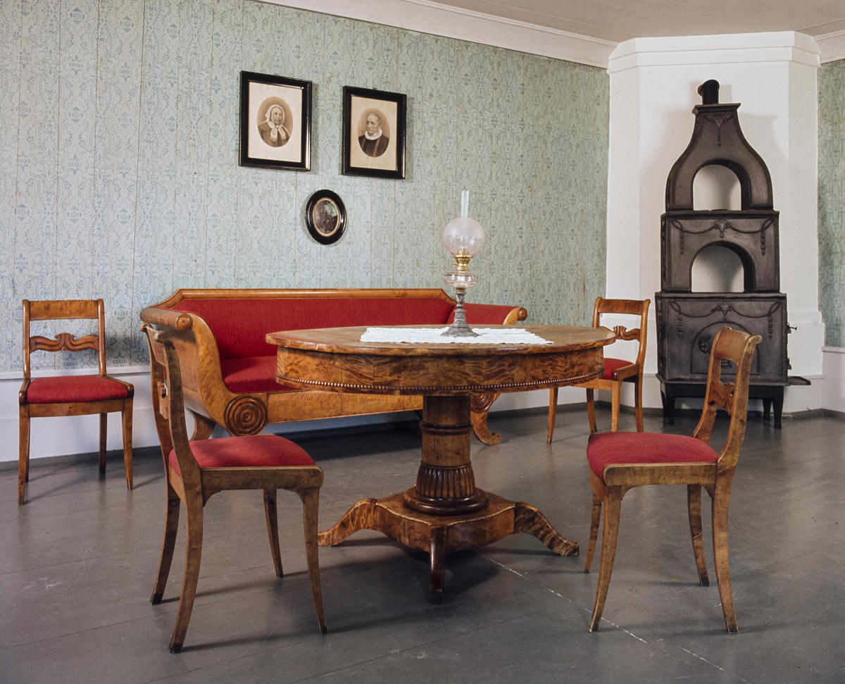Interiør; brunt bord med stoler og sofa i samme treverk og med rødt trekk, to gamle fotografier på veggen og en etasjeovn i støpejern.