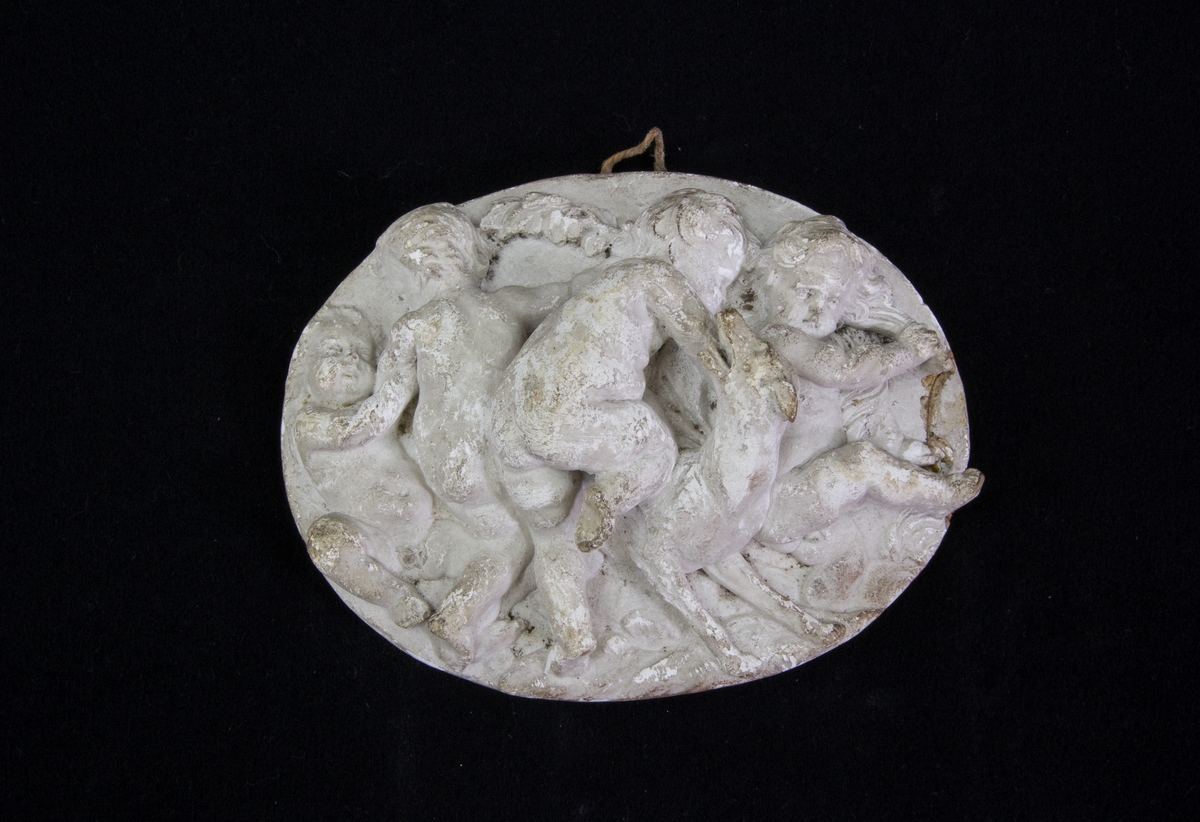 Reliefmedaljong av gips, oval, gråtonad. Föreställer fyra figurer likande putti samt hund. Snöre för upphängning.