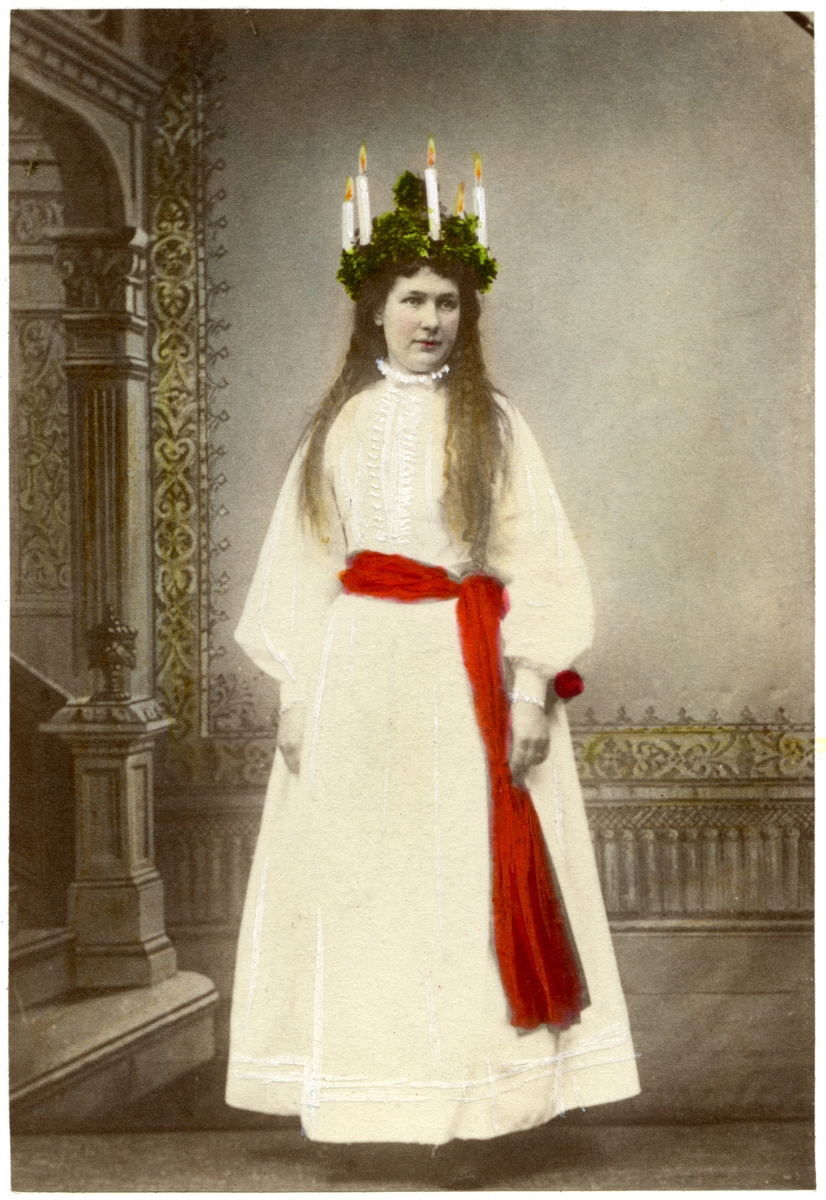Ateljéporträtt, kolorerat, föreställande Kröök Karin Olsdotter från Leksand i Dalarna som Lucia. Hon bär långt, vitt, linne, rött band i midjan och en klädd krona med ljus. Skansens Lucia omkring 1898.