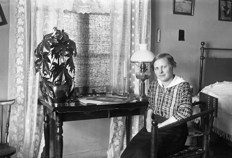 En kvinna sitter i en stol med ett bord bredvid sig.
På det finns en blomma och en lampa.
I bakgrunden syns en säng.
Agda Fors (1888-1964), blivande fru till fotografen.