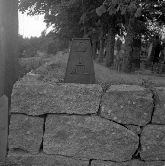 Foto av en milsten av järn. insatt i muren kring en kyrkogård. 
Text: "1 Mil." 
I SÖ hörnet av kyrkogårdsmuren till Älmeboda kyrka. 
Vägen Linneryd-Yxnanäs. 
Källa: Kronobergs läns väginventering 1943.