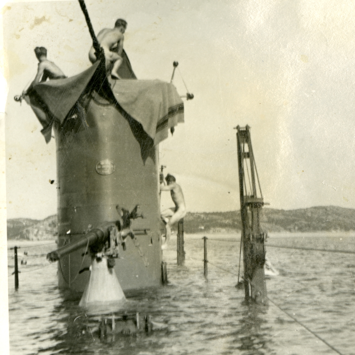 Bilden visar ubåten Najad vars däck ligger under vatten, bara tornet och överbyggnader sticker ut. På tornet syns tre nakna ubåts soldater, den ena håller på att klättra upp på sida av tornet. Runt omkring i vattnet syns fler besättnings män som badar.