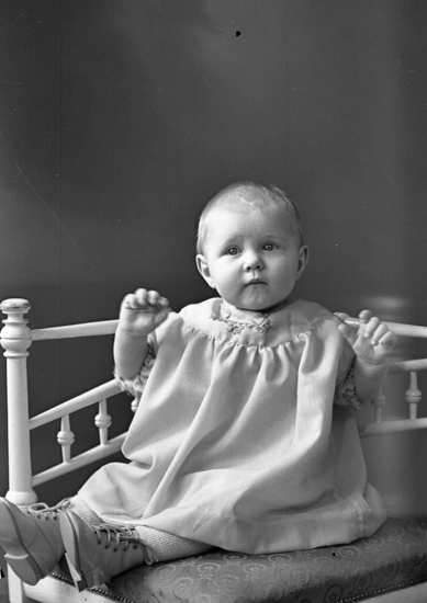 Foto av en okänd liten flicka i klänning. Hon sitter i en vitmålad stol.
Helfigur. Ateljéfoto.