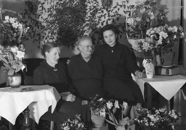 Foto av en äldre dam i glasögon och klänning, som sitter i en soffa i ett vardagsrum, med dottern Vivi Kristina (?) och ? .
Trol. fru Anna Helena Andersson (3 mars 1884-1979), Sållareboda, Hjortsberga, 
Källa: Sveriges Dödbok 1901-2009.