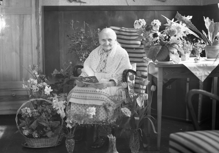 Foto av en okänd äldre kvinna i klänning och sjal. Hon sitter i en gungstol i ett dagrum, omgiven av blommor.