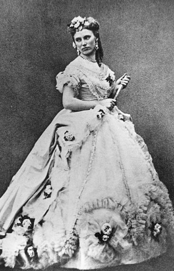 Rollporträtt (?) av Christina Nilsson. Hon bär en stor klänning och en solfjäder i ena handen. Hon har även blommor i håret.
Violetta i La Traviata.