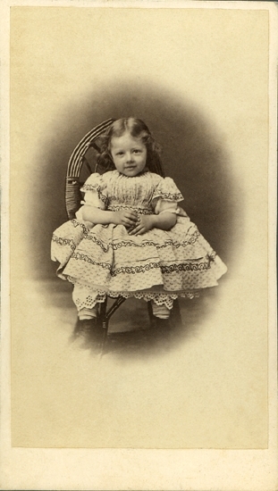 Porträtt (helfigur, en face, oval) av en sittande flicka i rutig klänning med rysch och volanger. Man skymtar också en spetsprydd underkjol, tjocka vita strumpor och ett par kängor. Hon sitter i en korgstol. 

Signe Christina Emilia Selldén (1862-1937) dotter t. fältläkaren Sven Niclas Selldén* (1806-1882) och hans 2:a hustru Mariana Erika Christina von Otter (1822-1869), Växjö.
G.1884 m. sedermera generallöjtnanten Pehr Richert Hasselrot (1861-1937), Stockholm. 
Källa: Svensk Släktkalender 1936. 
* Gift 1:a gången med Emilie Ulfsax (1812-1858).