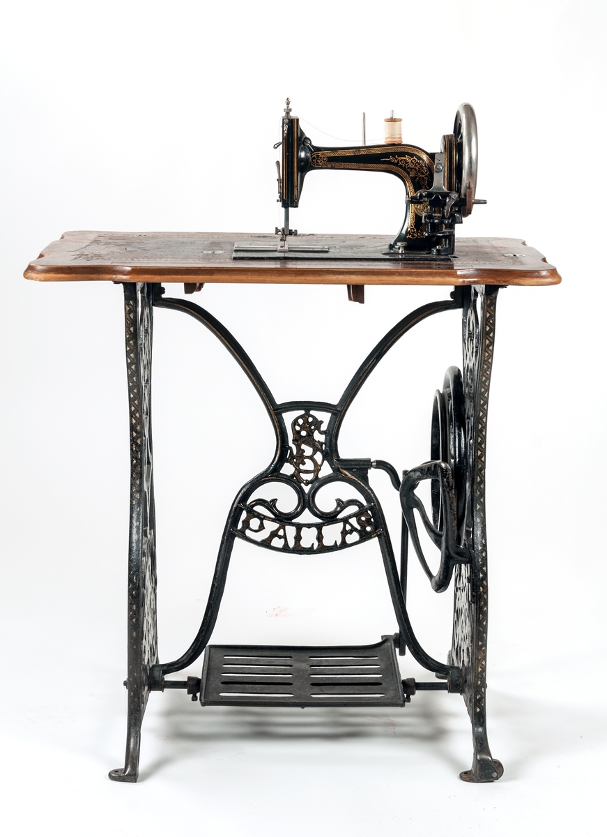 Symaskin av märket Pallas, tysk modell. Trampmaskin av gjutjärn med bord och huv av trä.
Maskinen har ett emblem med ett "S" och med ett "B" inne i maskinen.