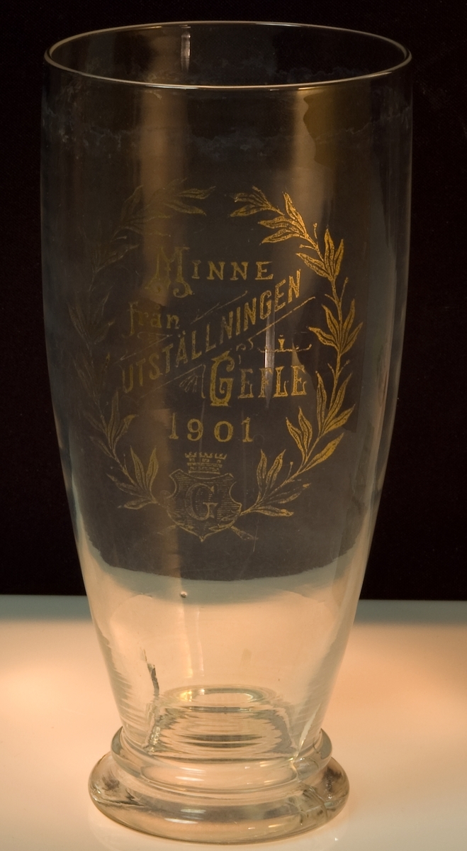 Dricksglas med text tillverkat för gävleutställningen 1901. Text på glaset: "Minne från utställningen i Gefle 1901" samt Gefle stads vapensköld. Texten något nött. Sprucken i foten.
