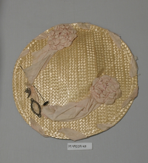 Platt gul flätad halmhatt med dekoration av rosa siden samt 
metallsmycken och en hattnål.
På undersidan fastsydd flätad halmring som är insnodd i rosa sidentyg
och en sydd rosa siden"ros".

Inskriven i huvudbok 2008-06-25