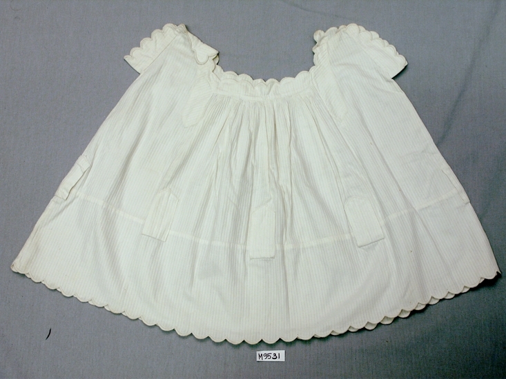 Barnklänning för barn, 2 års ålder.
Av vitrandigt bomullstyg, randen är ett vävt 
mönster vitt på vitt. 
Fyrkantig halsringning.
Urringning, ärmar och nedkant har uddavslutning 
med langettsöm.
Nedanför midjan åtta stycken klaffar avsedda för
sidenband.

Inskrivet i huvudbok 1937.
Funktion: Barnklänning