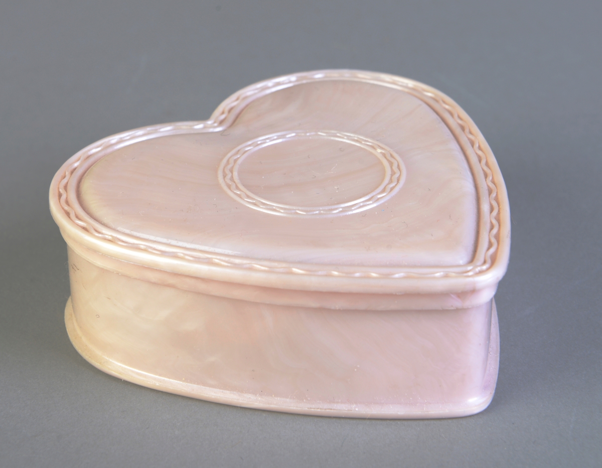 Et lite smykkeskrin laget av rosa plast. Skrinet er formet som et hjerte og lokket er dekorert med bord langs kanten og i en sirkel på midten. I skrinet ligger det et armbånd og diverse nåler/brosjer.