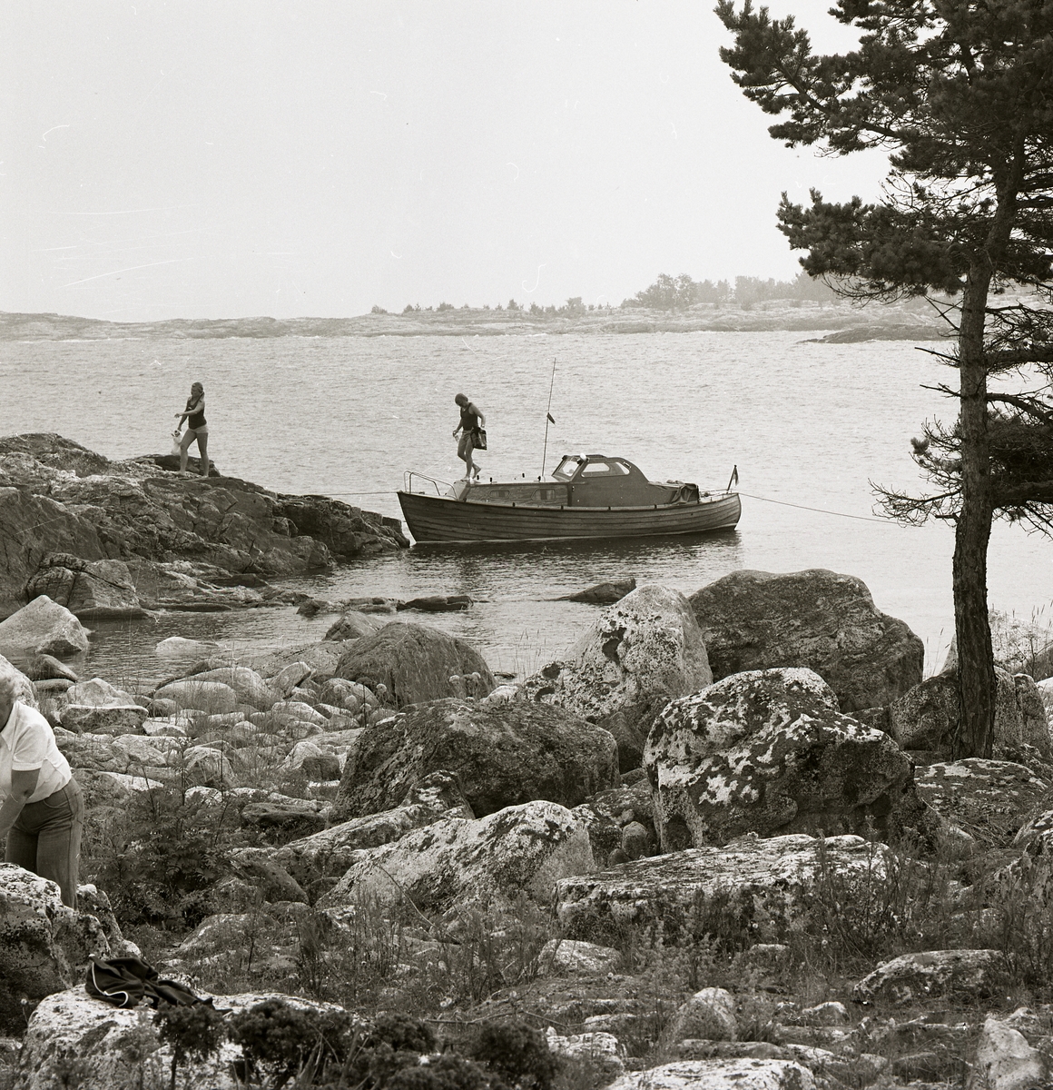 Invid en stenhäll i Gästriklands skärgård har en båt lagt till och två personer kliver iland. I förgrunden står en tall bredvid en stenröse och en tredje person skymtar förbi i vänstra hörnet. Bakgrunden fylls av havet och närliggande stenhällar.
