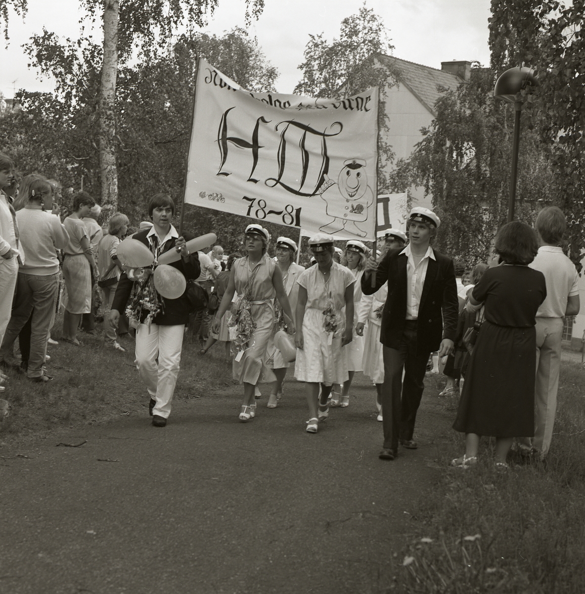 Studenter tågar bakom banderoller upp för Ohnbacken, Bollnäs den 4 juni 1981. Längs vägkanten har en publik ansamlats för att bevittna studenttåget. Alla studenter bär vita studentmössor och har runt sina halsar blommor och ballonger. På banderollerna går att läsa klassernas namn, studieperiod och i vissa fall är även en humoristisk komponent tillagd.
