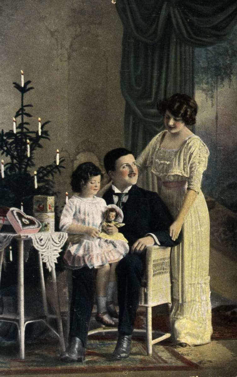 Julekort. Jule- og nyttårshilsen. Kolorert fotografi. Mor, far og datter ved juletreet. Datteren med dukke. Julegaver. Datert 1919.