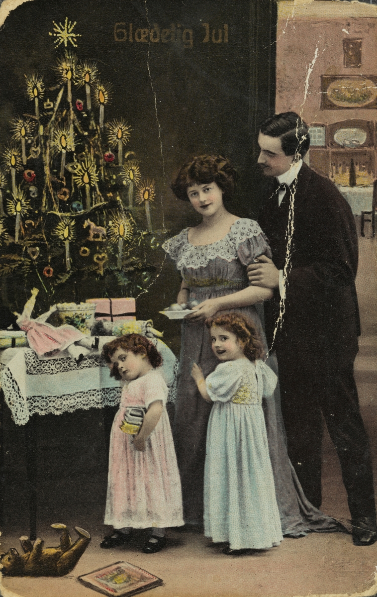 Julekort. Jule-og nyttårshilsen. Ant. Familie med to piker foran et juletre med gaver under. Håndkolorert fotografi. Utenlandsk kort.