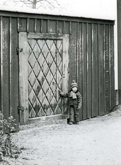 En pojke står intill en dörr i ett plank. Pojken bär vinterkläder.