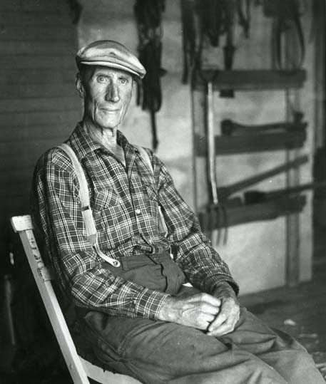 En man med arbetsbyxor, rutig skjorta och keps sitter med händerna i knät. Han är modell åt skulptören Per Nilsson-Öst i hans verkstad.