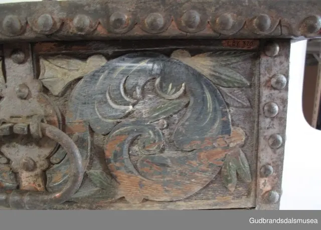 "Bladformer, smøygde band og drageornamentikk. Millom kantjarn og utskurden hev lokket fint utskorne små blad og halvrunde aggar. Vel vedlikehalde, sterk jarning og prydelege gangjarn. Truleg frå 1500 talet. Fra Haugen, Øystre Gausdal". (Jf. "Katalog for Lom Bygdesamling,1925).
Skrinrammen er holdt sammen av utvendige hjørnekantbeslag; jernhandtak i hver side. Fronten har nøkkelhullslås med beslag rundt. Lokket er satt samen av 2 bord, forsterket med jernbeslag rundt kanter på overside; to hengselbeslag festet til innside av lokk og bakstykke. Skrinsider blir holdt sammen av beslag ned langs hjørnekanter; har jernhandtak festet på jernbeslag.
Bunnen er satt sammen av 2 bord, festet til hverandre med to forsenka treplugger; bunnen var opprinnelig festet til underkant av skrinsider med 4 x 4 trenagler,seinere festet med spiker. 
Det er jernbeslag  på bakstykket midt, rundt hjørnekanter, omkring nøkkelhullslås, rundt lokk samt 2 handtak med beslag. Alle beslag er festet med store jernnagler; beslag på sidestykker er festet med små jernnagler under skrinbunnen. 3 av 8 bunnfester for beslag er fortsatt intakte.
DEKOR. De store jernnaglene med forskjøvet plassering på beslag ble sannsynligvis brukt som dekorelement; runde beslag på sider, bakstykke og front. Flatskurd på lokk med flettet, stilisert renessanseranke, der bladverk er "omgjort" til småspiraler eller rundinger.  På skrinsidene akantusskurd med c-former, bladtunger uten stilk og naturalistiske småblad i rokokkostil. Flatskurd var vanlig i Gudbrandsdalen i perioden ca. 1650-1750, mens akantusmotivet først ble vanlig på bruksgjenstander i løpet av siste halvdel av 1700-tallet. Den mørkere grønne og blå dekorfargen var vanlig i siste del på 1700-tallet; den brune bunnfargen var vanlig i bruk fra 1780-tallet. 
BRUK. Før kofferten kom i bruk hadde man reiseskrinet, som ble brukt til oppbevaring av bl.a. klær, verdisaker og mat på reiser. Skrinet hadde ofte et eller to mindre, smale rom langs den ene skrinsiden og ett større rom. De fleste reiseskrin var i tillegg forsterket med jernbeslag langs alle kanter og med en solid nøkkelhullås til sikring mot støt, slag og tyveri.