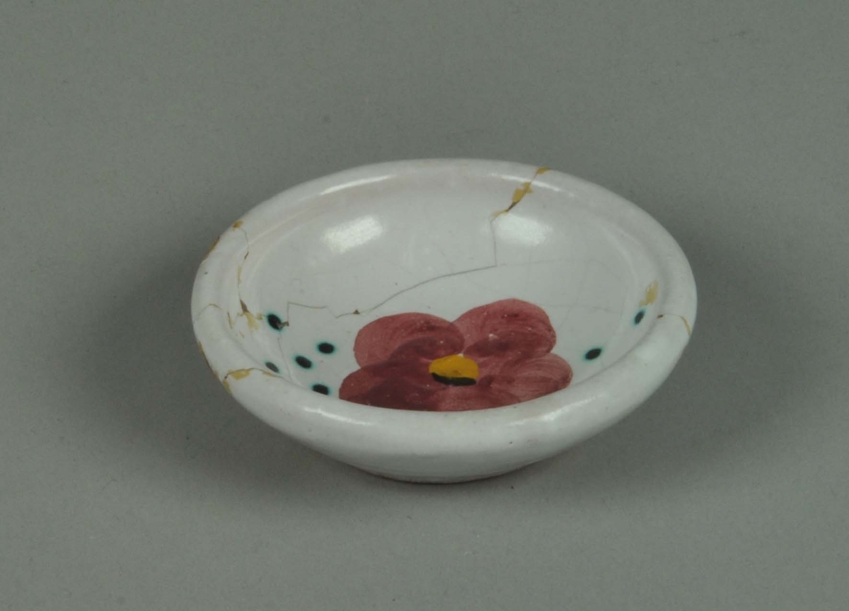 Keramikkskål med hvit glasur og påmalt blomstermotive og grønne prikker.Skålen er limt.