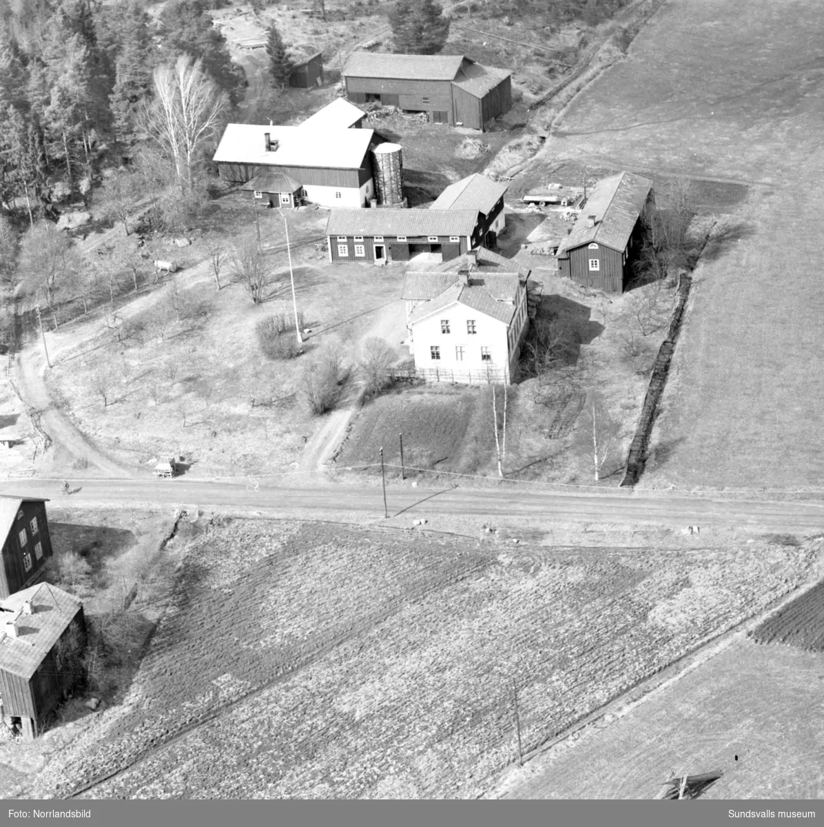 Flygfoto över en oidentifierad gård, troligen någonstans i Selånger. På bild två har helikoptern landat på en lägda och människor har samlats runt den.
