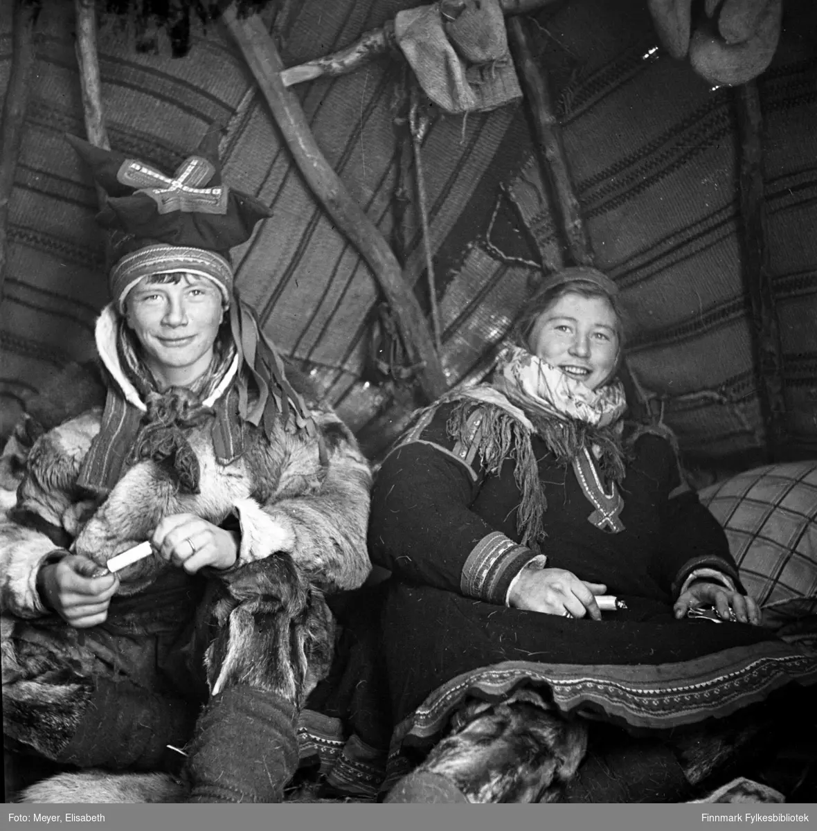 Søskenparet Mathis Johansen Sara og Inga Johansdatter Sara fotografert inne i lavvoen. Fotografert i Kautokeino i periden 1939-40. Begge er kledd i samiske drakter. Mathis bærer samisk lue, pesk og har skaller og skallebånd på beina. Inga har samisk kofte og lue med sjal i halsen. Et liknende bilde ble publisert i Nordmannsforbundets julehefte 1950 og Magasinet for alle nr. 13, 29.mars 1961.
