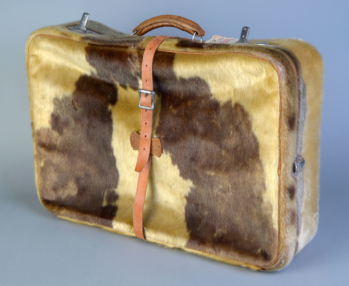 Koffert laget av selskinn med pelsen ut. Den er mørk brun og gulhvit på farge. I fronten er det to koffertlåser på hver side, begge med nøkkelhull. På sidene er det en enkel lås på hver side. Det er et skinnbelte med beltespenne rundt kofferten. Håndtaket er i fronten og er laget av skinn og festet med to metallringer i skinnfester på kofferten. Kofferten er trukket med rødbrun silkemoare inne, både i bunn og lokk. I bunnen er det to lærreimer med spenner for å holde tøyet på plass. Bakerst er det en lomme i rødbrun silkemoare med strikk i. På hanken er det festet en lapp fra SAS.
