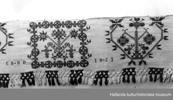 Hängkläde av linne med röda hallandssömsbroderier. Vita bomullsband i form av hällor är fastsydda i överkanten. Märkning, broderat: "EBBD", "1853". Frans +:70 mm. 