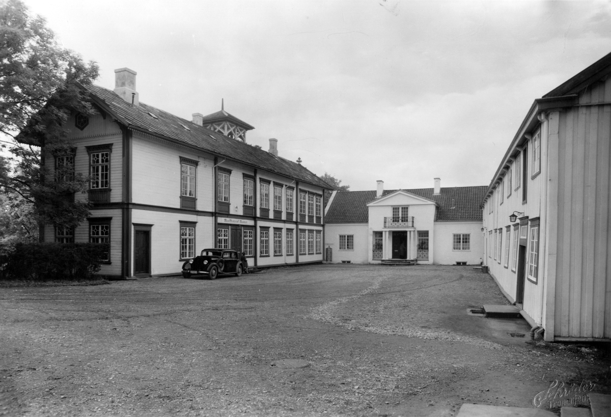 Bilde av tunet med hovedbygningen til venstre, direktørboligen i midten og Wesselbygningen til høyre. Det står en bil parkert fremfor hovedbygningen.